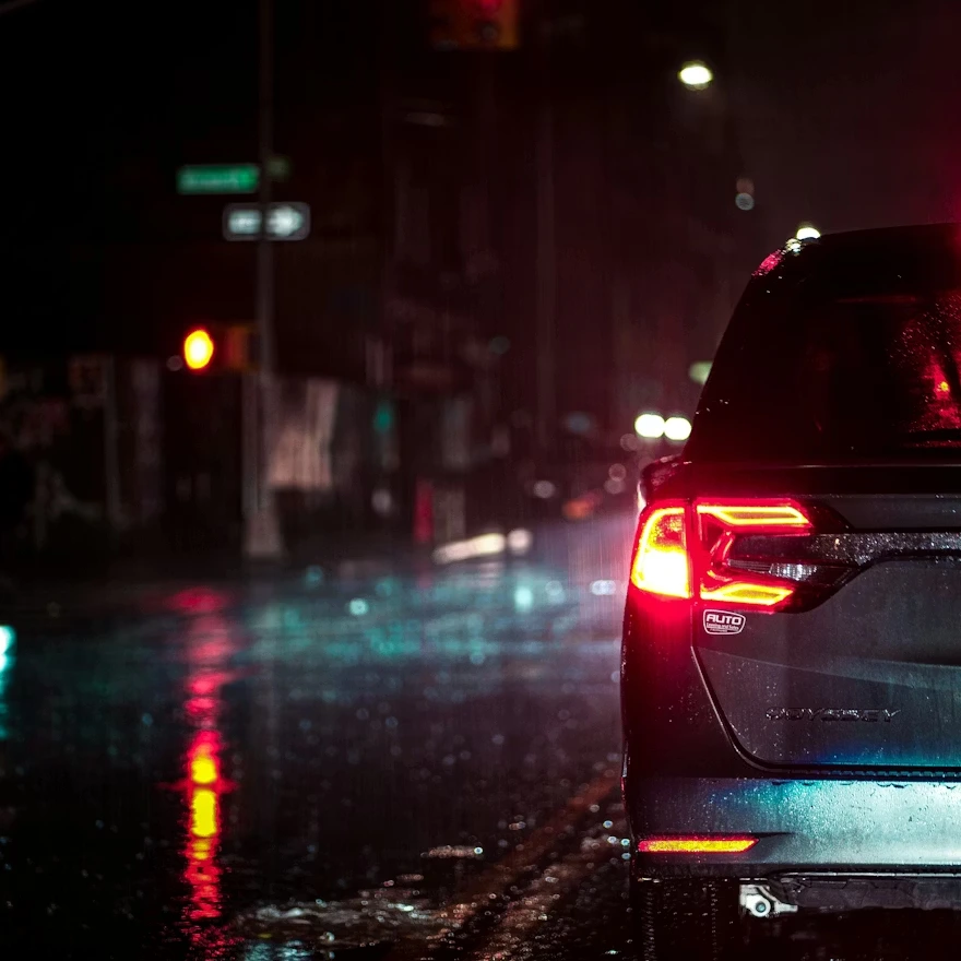 Automóvil detenido de noche bajo la lluvia, con las luces traseras encendidas reflejándose en el asfalto mojado, en una calle urbana con semáforos y luces difusas al fondo