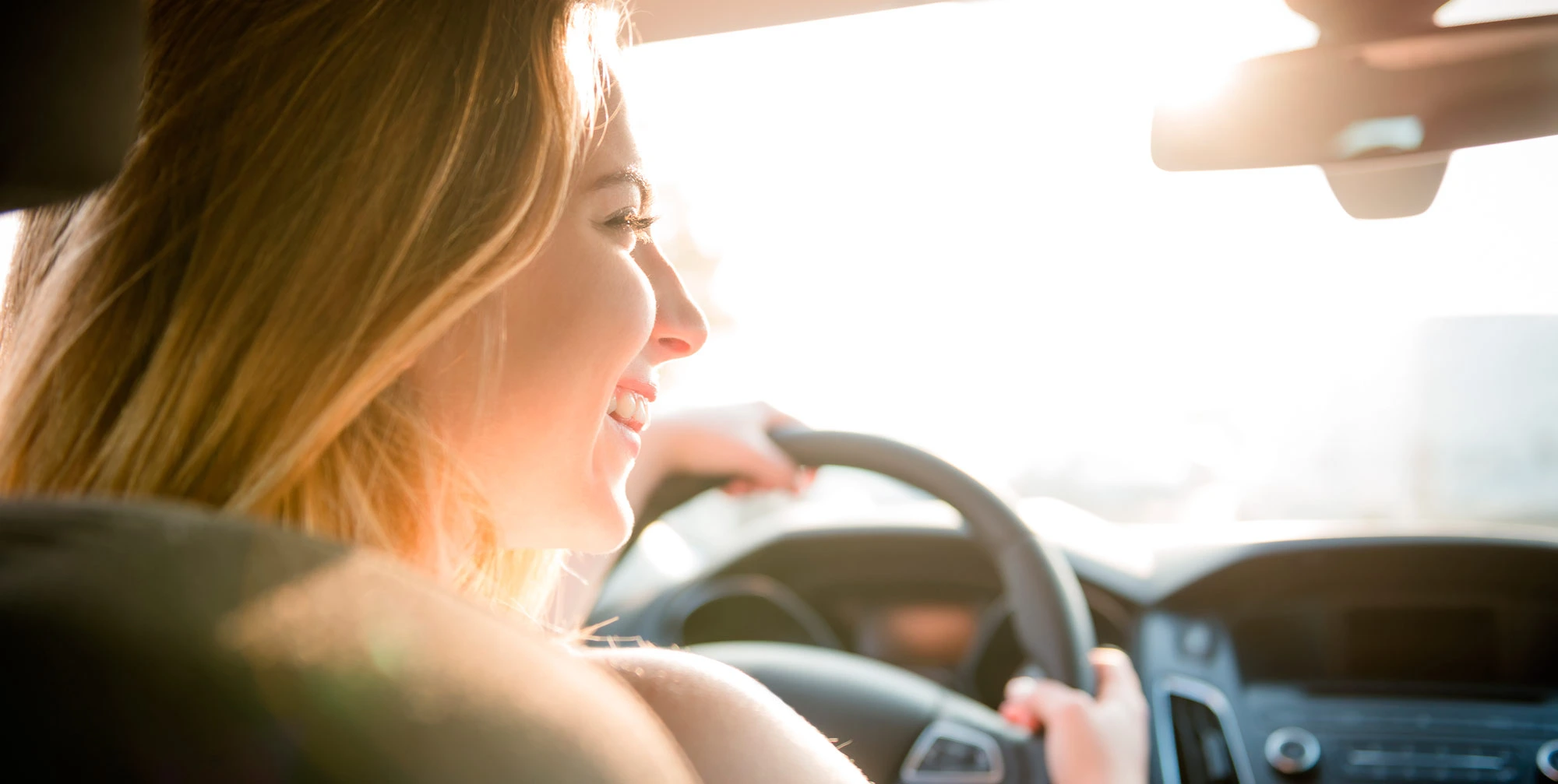 Primer plano de una mujer conduciendo un automóvil, vista de perfil desde el asiento trasero, sonriendo mientras sostiene el volante con luz cálida del sol entrando por el parabrisas.