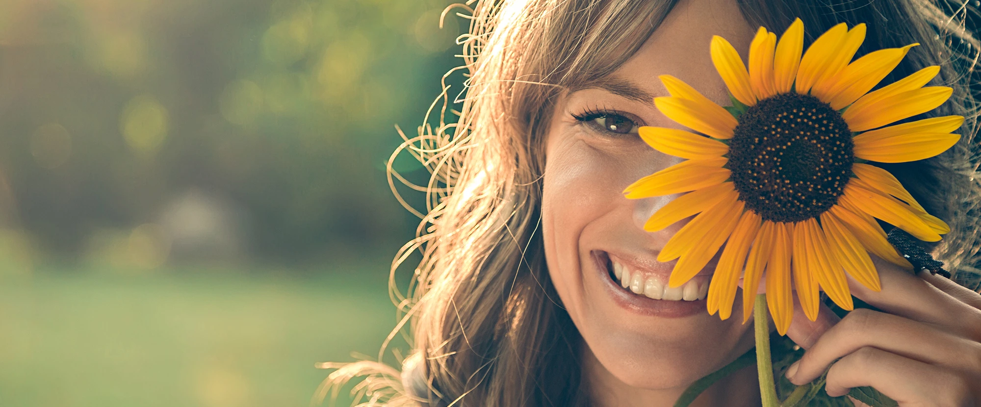 Primer plano de una mujer sonriendo al aire libre, sosteniendo un girasol amarillo.